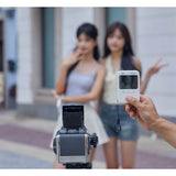 A hand holds a Godox C100 Transparent Viewfinder Camera in the foreground, while a camera and two blurred people appear in the background in an outdoor setting.