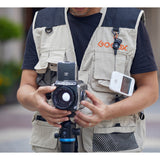 A person wearing a beige and navy Godox photography vest holds a medium format film camera with a viewfinder and attached accessories, including a Godox C100 Transparent Viewfinder Camera in an outdoor setting.