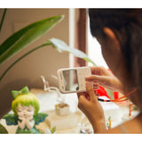 A person holding a Godox C100 Transparent Viewfinder Camera, shown in a lifestyle setting with a blurred background and a decorative figurine.