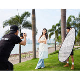 A Godox W-RFT130 130cm (52") Windproof Silver Collapsible Reflector being held by an assistant while a photographer takes photos of a model in a park