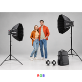 Two people standing between two Godox studio softbox lights in a studio setup, with a black Godox backpack and two small black-and-white checkered flags on the floor. The word 'RGB' appears in multicolor text at the bottom center.