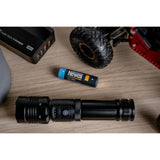 The Newell 18650 USB-C Rechargeable battery with a Black torch, and a tyre from an RC car on a wooden surface with a blurred background