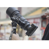A black mirrorless camera body with an attached telephoto lens and a mounting bracket, held by a person in an outdoor setting with a blurred background.